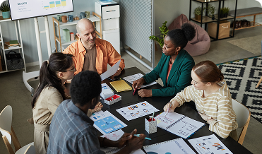 A group of diverse coworkers sit at a table and discuss partner marketing strategy over reports and graphs